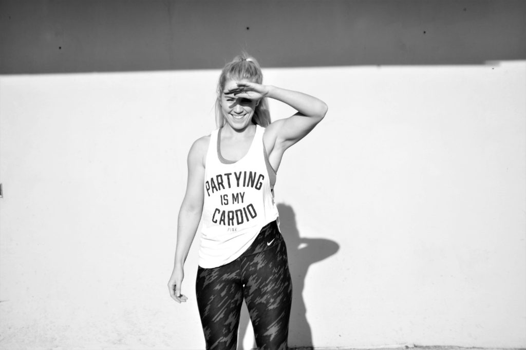 In this post you will learn - 80+ Helpful Fitness Terms To Know As A Beginner. 
This picture is of Jane wearing a t shirt saying 'Partying Is My Cardio, with patterned Nike leggings. She is covering her eyes from the sun.