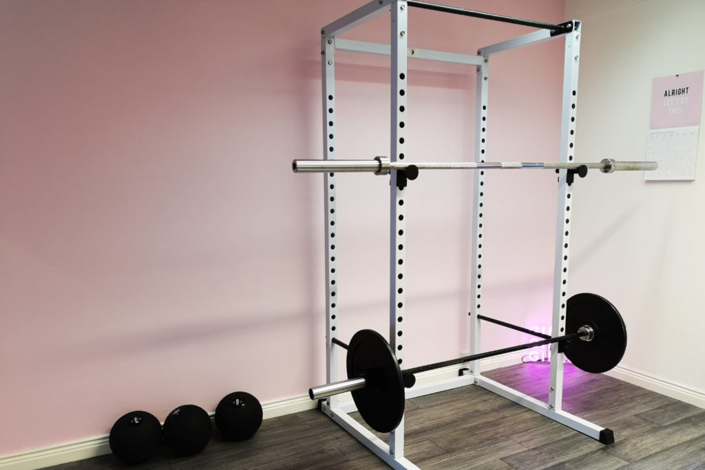 In this post you will learn - 80+ Helpful Fitness Terms To Know As A Beginner. 
This picture is of a white squat rack, with two barbells resting on it. There are 3 medicine balls resting against a pink wall, with a pink calendar on it saying 'Alright, Let's Do This.'