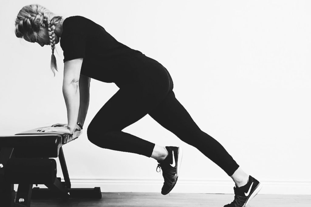 In this post you will learn - 80+ Helpful Fitness Terms To Know As A Beginner. 
This picture is of Jane wearing a black t-shirt, trainers and leggings, performing an inclined mountain climber exercise on a bench.